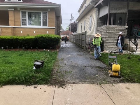 Workers operating equipment in residential alley between two homes on wet ground with green lawns visible