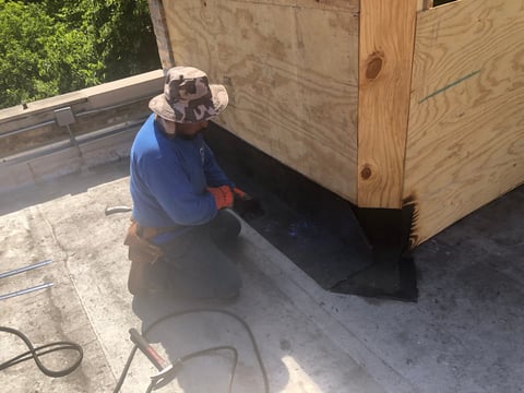 Worker in blue shirt applying black roofing material to flat roof surface with wooden framing structure