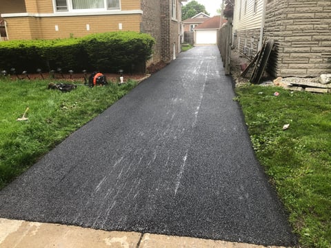 Newly installed dark asphalt driveway between residential houses with green lawns on either side