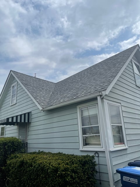 Light blue residential house with gray shingled roof, white trim windows, and green shrubs under cloudy sky